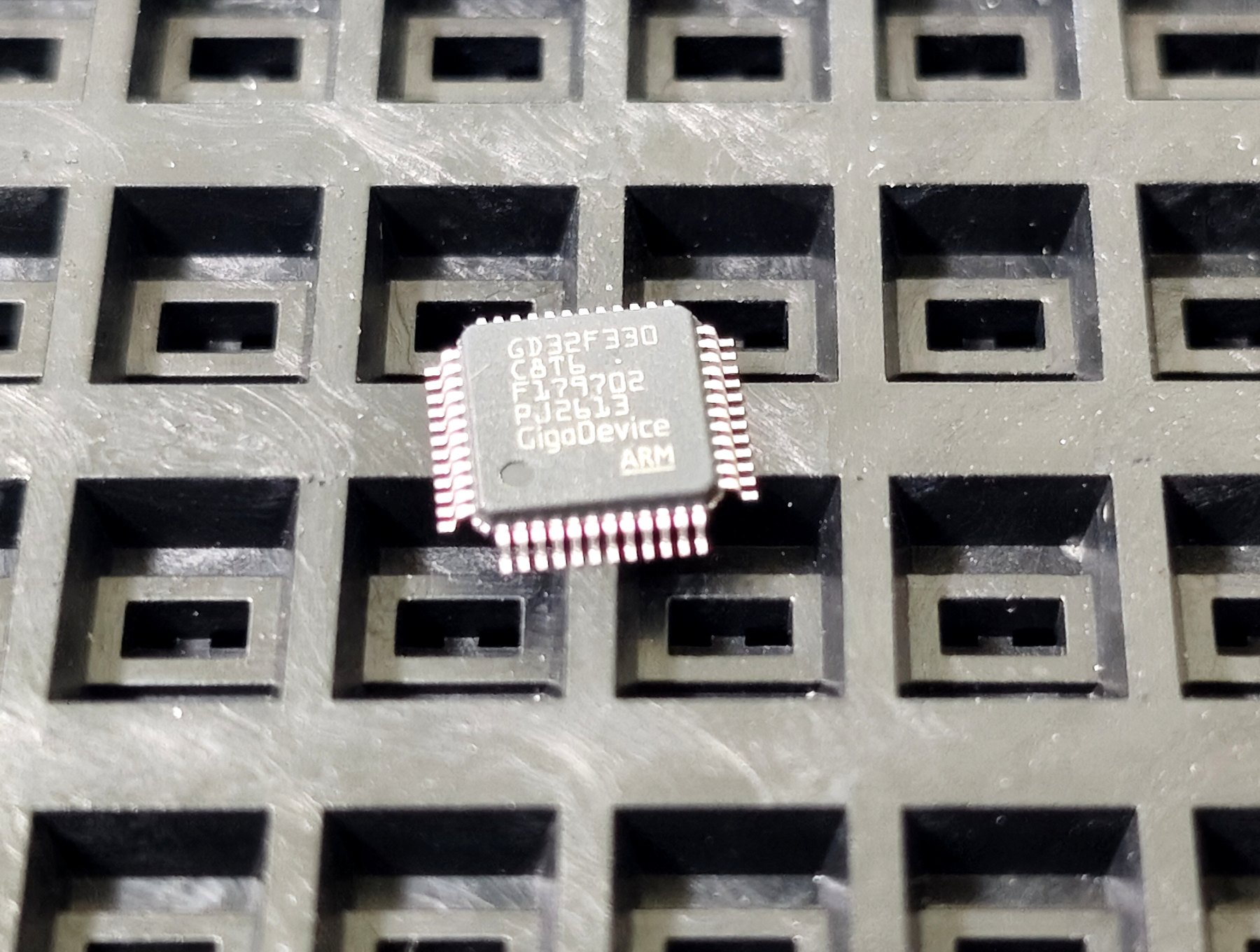 Close-up of an LQFP microcontroller in a waffle tray, with the manufacturer name, full part number, lot code and date code legible.